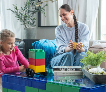 A young girl playing with Biggo Blocks, assembling a vehicle while her mother watches and smiles from the couch, showcasing a creative and playful moment with extra large building blocks.