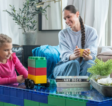 A young girl playing with Biggo Blocks, assembling a vehicle while her mother watches and smiles from the couch, showcasing a creative and playful moment with extra large building blocks.