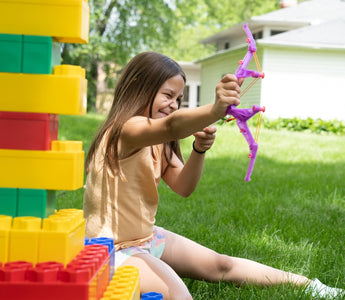 A young girl playing outdoors with colorful Biggo Blocks, aiming a toy bow and arrow, surrounded by green grass and a sunny background.