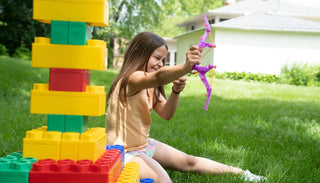 A young girl playing outdoors with colorful Biggo Blocks, aiming a toy bow and arrow, surrounded by green grass and a sunny background.