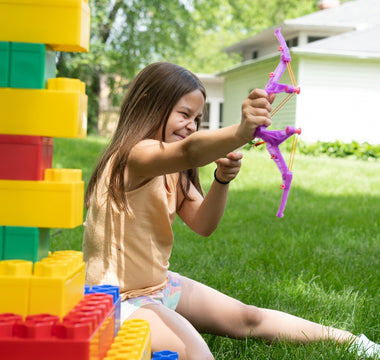 A young girl playing outdoors with colorful Biggo Blocks, aiming a toy bow and arrow, surrounded by green grass and a sunny background.