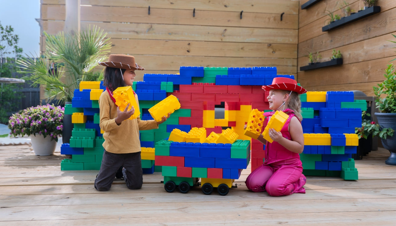 Two children dressed as cowboys play together with Biggo Blocks in a colorful backyard fort, smiling and holding jumbo building blocks.