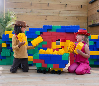 Two children dressed as cowboys play together with Biggo Blocks in a colorful backyard fort, smiling and holding jumbo building blocks.