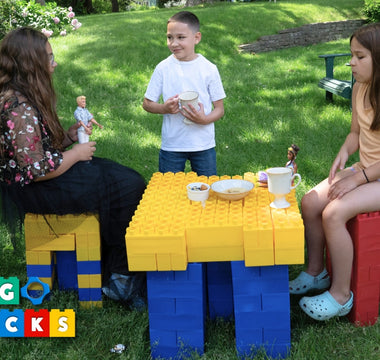 Siblings having a tea party outside using Biggo Blocks to build colorful chairs and a table, surrounded by grass and sunshine.